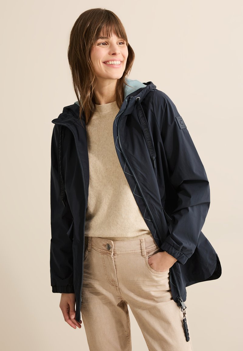 Chaqueta impermeable de color azul marino con cremallera frontal, capucha ajustable, forro contrastante en azul claro y puños elásticos, combinada con un suéter beige y pantalones.