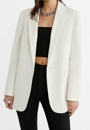 Femme portant un blazer blanc à un seul bouton, un crop top noir, un pantalon noir taille haute et un gros collier chaîne en argent.