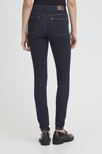 Osoba stojící zády, oblečené v tmavě modrých úzkých džínách s zadními kapsami a černých kožených mokasínách na světlém pozadí.