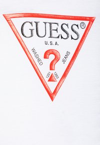 Weißes T-Shirt mit einem roten dreieckigen Logo und schwarzem Text, auf dem "GUESS U.S.A." und "WASHED JEANS" steht, mit einem Fragezeichen-Design.