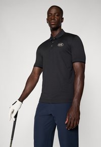 Polo shirt noir avec une texture subtile et une patch de marque, associé à un pantalon bleu marine et un gant de golf, tenant un club de golf.
