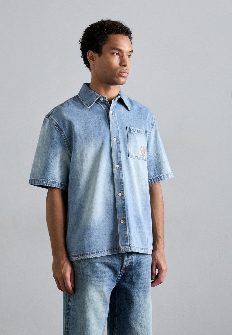 Lys blå, kortermet denimskjorte med brystlomme og knappeåpning. Falmet gradientdesign, med lett tekstur synlig på stoffet.