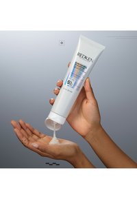 Redken Acidic Bonding Concentrate in einer weißen Tube mit Flip-Deckel. Ein klar-cremiges Produkt wird auf eine Hand gegen einen grauen Hintergrund ausgegeben.