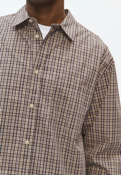 Homme portant une chemise à carreaux beige et marron boutonnée sur un T-shirt blanc, visible du cou jusqu'au milieu du torse sur fond blanc.