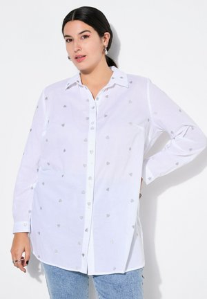 Chemise blanche à boutons avec broderie en forme de cœur argenté, manches longues, col et coupe décontractée. Portée avec un jean en denim bleu clair.