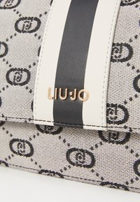 Bežna torba s crnim tekstilnim uzorkom. Ima crnu prugu i zlatni detalj s logotipom. Tekstura tkanine dodaje dubinu dizajnu.