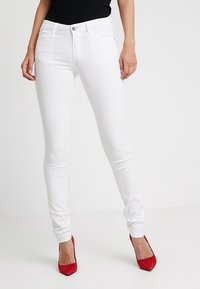 Jean skinny blanc en denim avec une texture lisse, présentant des poches avant et un design classique à cinq poches, accompagnés de talons rouges.