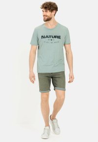 Lichtgroene t-shirt met de tekst "NATURE IS ALL WE NEED", gecombineerd met opgerolde olijfgroene shorts. Witte sneakers maken de outfit compleet.