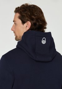Sudadera azul marino con una textura suave, con una capucha grande y un detalle del logo en blanco en la parte posterior. Diseño simple y minimalista.
