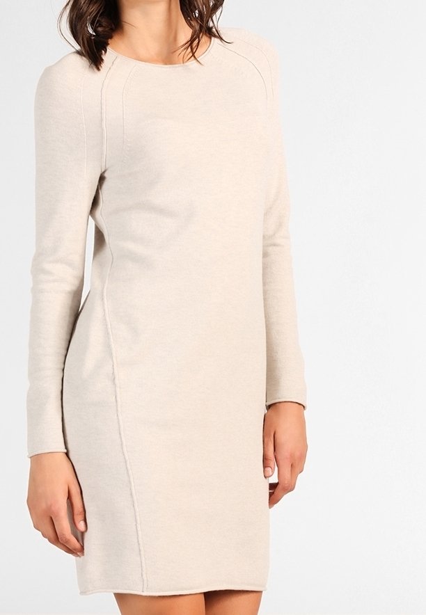 Modella che indossa un vestito lungo a maniche lunghe beige chiaro, in piedi davanti a uno sfondo semplice, con una mano posizionata vicino alla vita.