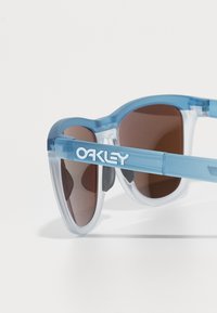 Kék áttetsző napszemüveg barna lencsékkel, kerek dizájnnal és feltűnő "OAKLEY" márkajelzéssel a szárán.