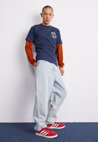 T-shirt bleu marine par-dessus un manches longues orange, jean ample bleu clair, et baskets rouges avec des rayures blanches. Texte sur le t-shirt : « PAIX & AMOUR ».