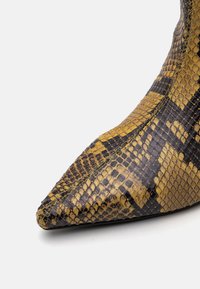 Szukane buty na spiczastym obcasie z sztucznego wężowego skóry w kolorze żółtym i czarnym. Posiadają teksturowaną powierzchnię z połyskiem oraz kontrastowe zielone przeszycia.
