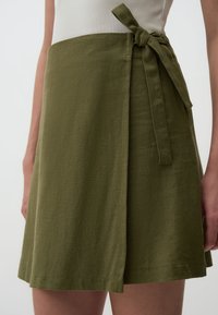 Jupe portefeuille vert olive en tissu texturé, avec une taille à nouer et une coupe droite, dotée d'une fente sur le côté.