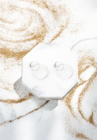 Silverfärgade hoopörhängen med en dubbel-loopdesign, visade på en oktagonal marmorstand, mot en bakgrund av guldglitter.