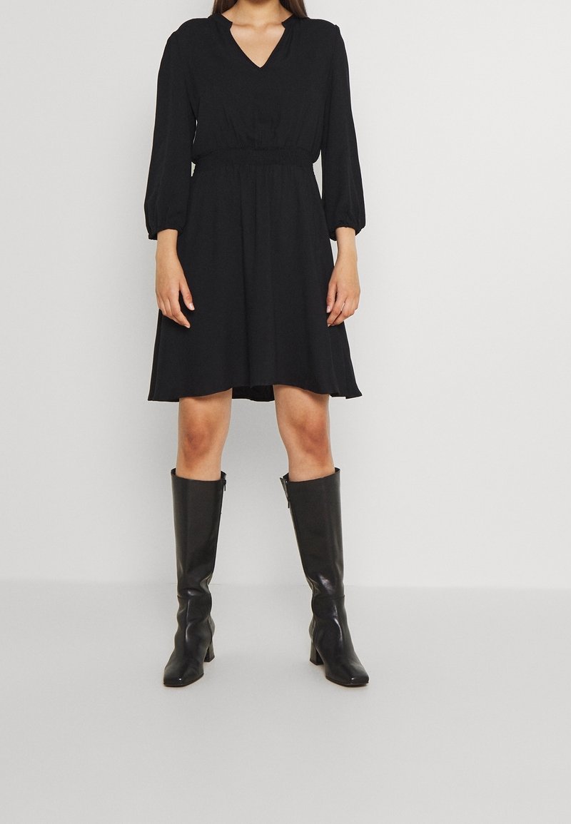 Svart, knälång klänning med V-ringning och långa puffärmar. Har en elastisk midja och en luftig siluett, tillsammans med svarta knähöga stövlar.