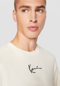 Jeune homme aux cheveux courts portant un t-shirt blanc à col rond avec une petite signature noire brodée sur la poitrine.