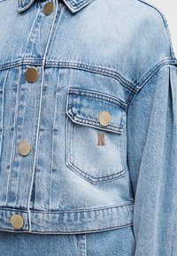 Ανοιχτό μπλε τζιν μπουφάν με μεταλλικά κουμπιά, εμπρόσθια τσέπη με καπάκι και ορατές ραφές.