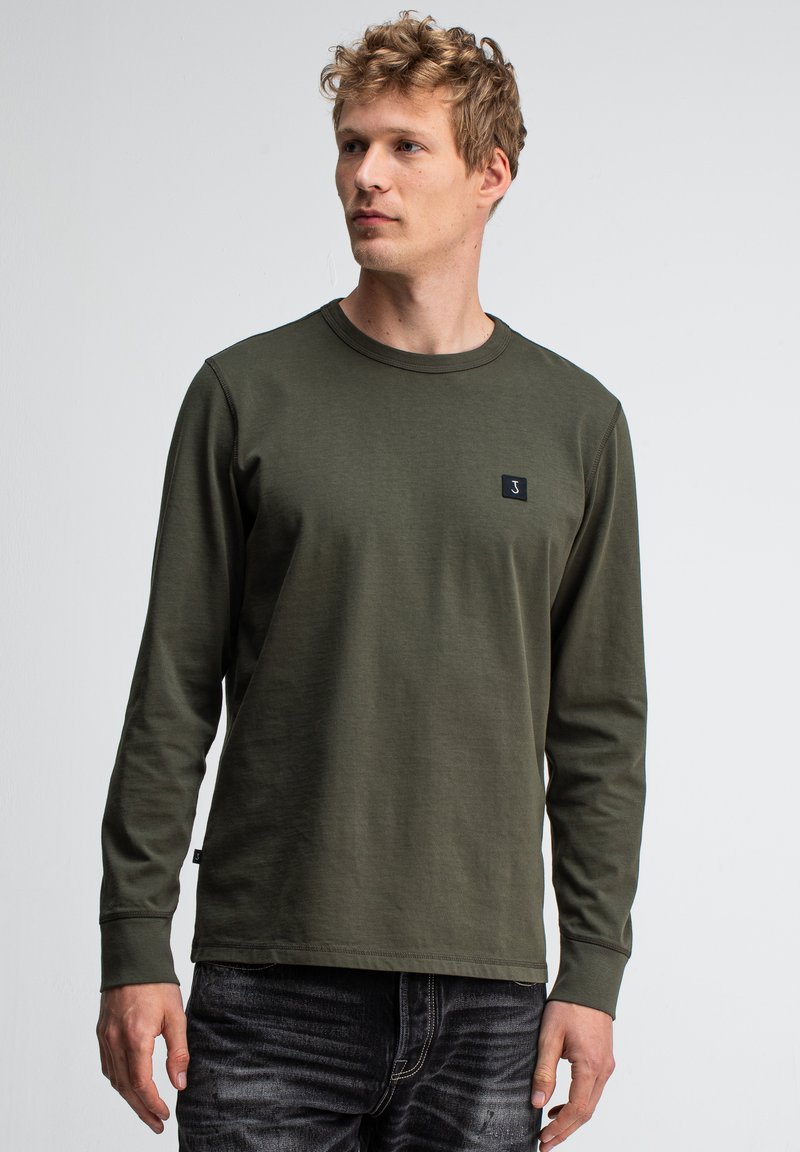 Junger Mann mit hellbraunen lockigen Haaren, der ein olivgrünes Langarmshirt und dunkel gewaschene Jeans trägt und seitlich vor einem schlichten Hintergrund schaut.