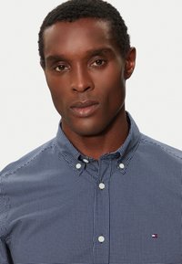 Hombre con cabello rizado corto que lleva puesta una camisa de botones de patrón azul marino con un pequeño logo bordado en el pecho.