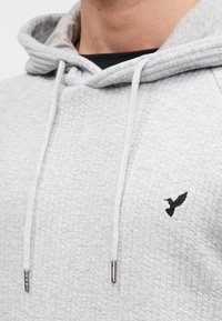 Ανοιχτό γκρι φούτερ με υφή υφάσματος, λευκά κορδόνια και μαύρο κεντητό λογότυπο πουλιού στο στήθος. Μαλακό και casual σχέδιο.