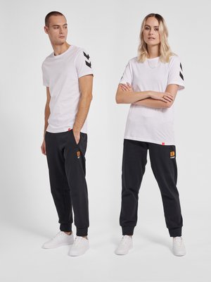 Mand og kvinde iført matchende hvide T-shirts med sorte chevronstriber på ærmerne, sorte joggers med orange logoer og hvide sneakers, stående side om side.