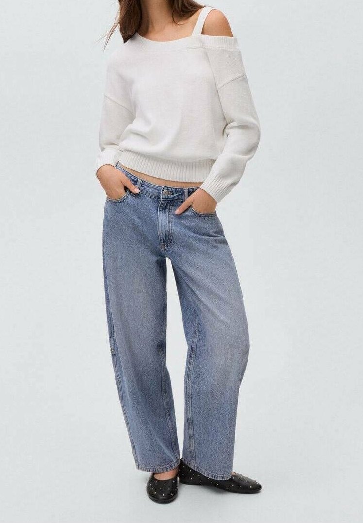 Femme portant un pull blanc épaules dénudées, un jean bleu décontracté et des ballerines noires cloutées, les mains dans les poches sur fond uni.
