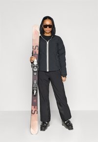 Skiudstyret, der vises, inkluderer en sort isoleret jakke med hvid lynlås, sorte bukser og skiger i pink nuancer med bindinger samt sorte støvler.