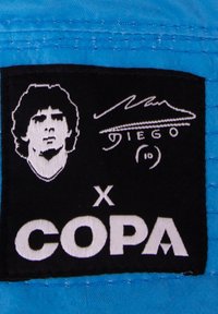 Zwarte stoffen patch met een witte geborduurde portret, handtekening en tekst "x COPA," bevestigd op een lichtblauw kledingstuk.