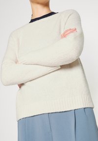 Personne portant un pull en maille crème à manches longues, les bras croisés sur la poitrine, associé à un pantalon bleu ample.