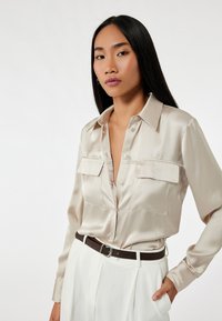 Camisa botón de satén beige con dos bolsillos en el pecho, combinada con pantalones blancos de cintura alta y un cinturón marrón oscuro. Textura suave, diseño clásico.