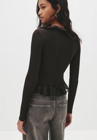 Top nero a maniche lunghe a coste con orlo a peplum realizzato in tessuto trasparente. Caratterizzato da un colletto arricciato e un design aderente. Abbinato a pantaloni in denim grigi.