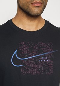 Camiseta de algodón negra con escote redondo. Presenta el logo de Nike en un diseño pixelado en azul y púrpura en la parte delantera. Textura de tela suave.
