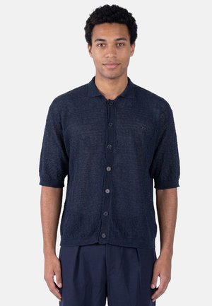 Uomo con capelli ricci che indossa una camicia blu navy a maniche corte con bottoni e pantaloni scuri, in piedi davanti a uno sfondo semplice.