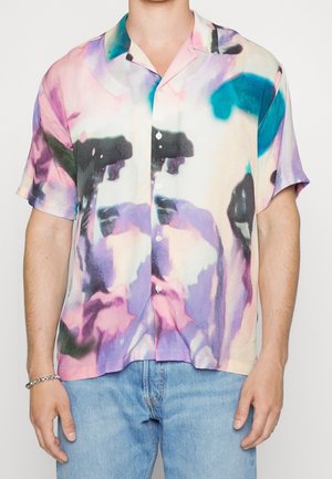Koszula z krótkim rękawem zapinana na guziki z abstrakcyjnym, pastelowym wzorem tie-dye w kolorach różowym, fioletowym, morskim i czarnym, noszona z jasnoniebieskimi dżinsami.