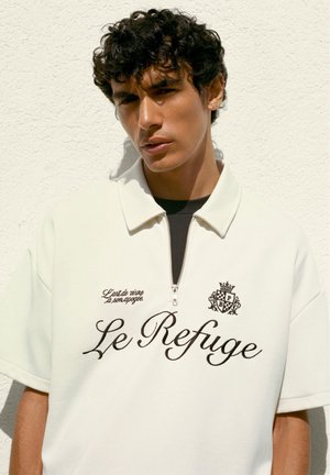 Jonge man met krullend haar, die een wit shirt met rits draagt met "Le Refuge" en een geborduurd wapenschild aan de voorkant, staand tegen een gestructureerde muur.