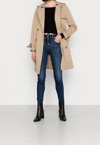 Trench beige con design doppiopetto e cintura, abbinato a jeans skinny blu scuro e stivaletti neri con tacco.