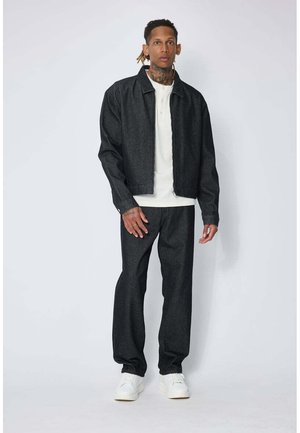 Homme avec des tatouages au cou portant une veste et un pantalon gris foncé, une chemise blanche et des baskets blanches, debout devant un fond clair uni.