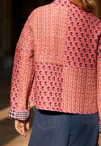 Femme portant une veste matelassée patchwork rose avec des motifs floraux et des manches retroussées sur un jean en denim bleu, vue de dos.