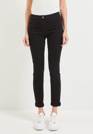 Jean noir slim avec revers roulés, porté avec des baskets blanches et un haut blanc sans manches, visible de la taille aux pieds.