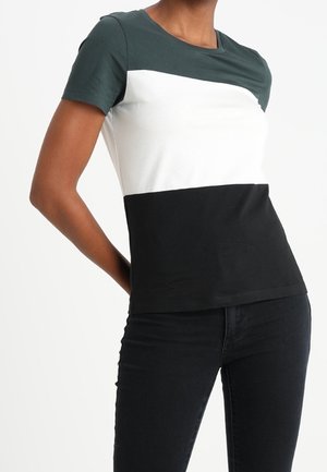 Kvinde iført en kortærmet t-shirt med farveblokke i mørkegrøn, hvid og sort horisontale sektioner, kombineret med sorte bukser.