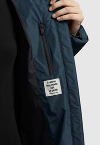 Interior de una chaqueta acolchada azul con una cremallera negra y una etiqueta que dice: "Un abrazo cálido hecho de tela" en texto blanco sobre la tela.