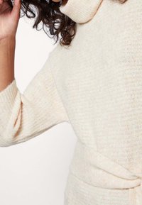 Personne portant un pull en tricot couleur crème avec un col haut et une ceinture, touchant ses cheveux d'une main.