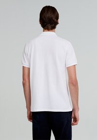 Camisa polo blanca de manga corta y cuello clásico, con una textura suave y dobladillo recto, combinada con pantalones oscuros.