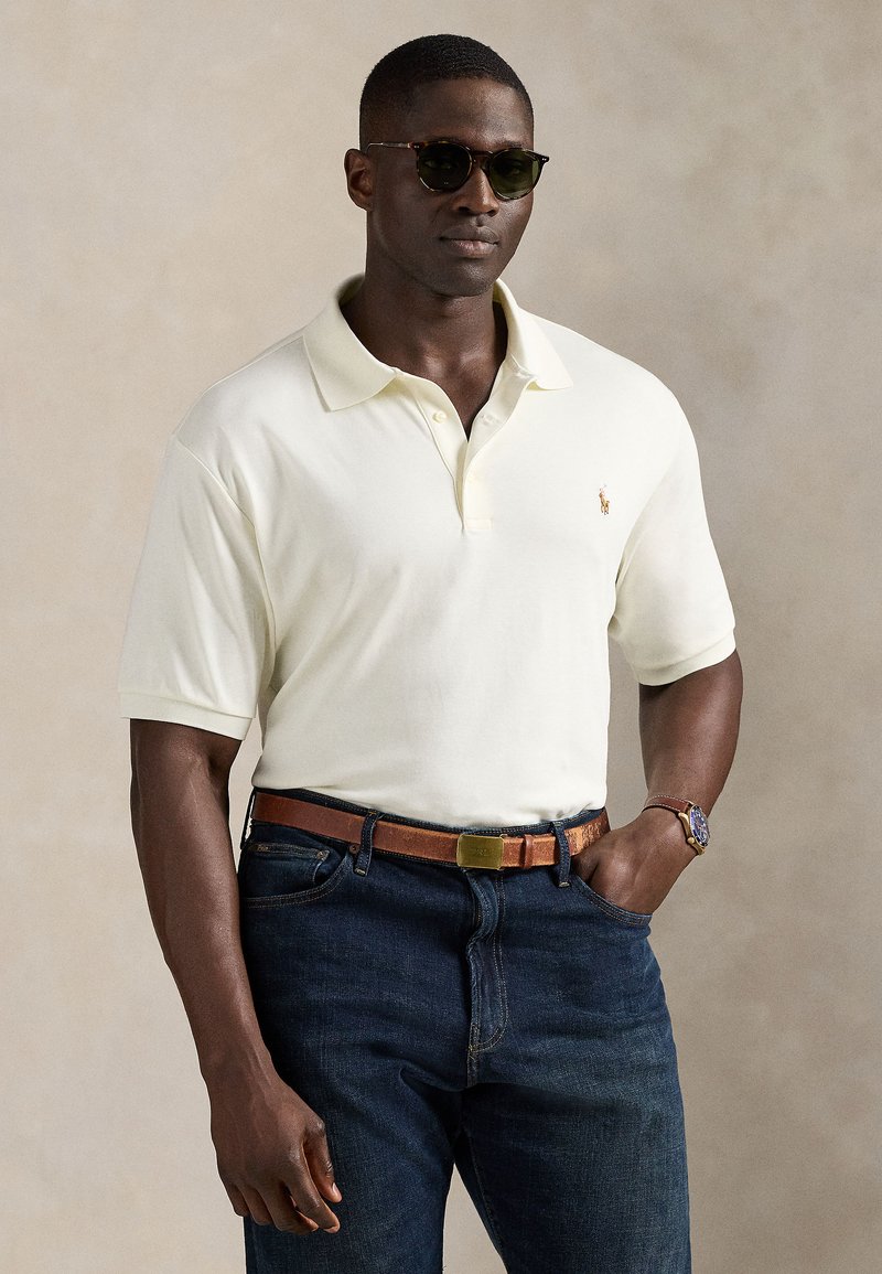 Lys gul poloskjorte med korte ermer, brodert logo, kombinert med mørkeblå denimjeans og et brunt skinnbelte. Tilbehør inkluderer solbriller.