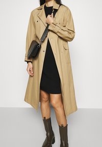 Cappotto trench beige con design doppiopetto, accessori in argento e tasche laterali, indossato sopra un vestito nero a coste e abbinato a stivali marroni.