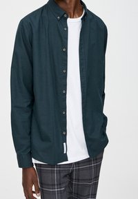Donkergroene button-up shirt van katoen, met een kraag en lange mouwen, gedragen over een wit T-shirt en grijze geruite broek.