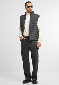 Schwarze gepolsterte Weste über einem cremefarbenen Rollkragenpullover, kombiniert mit dunkelgrauen Cargo-Hosen und schwarzen klobigen Stiefeln. Die Accessoires umfassen Sonnenbrillen.