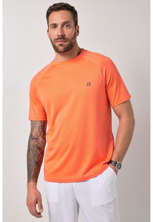 Mann trägt ein helloranges Kurzarmhemd mit kleinem Logo, weiße Hose und eine Armbanduhr, steht mit einer Hand in der Tasche.
