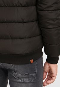 Crna puffer jakna s kvadratnim uzorkom, rebrastim rubom i smeškom kožnim logotipom; uparena s izblijedjelim sivim trapericama koje prikazuju detalje džepova.